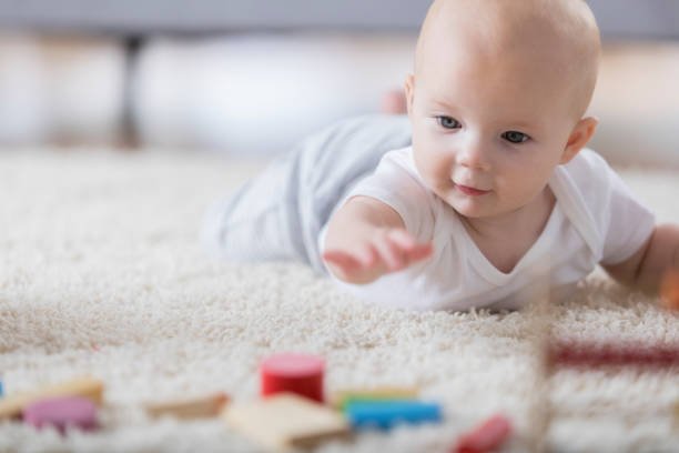 Focus is on an adorable baby laying on a rug in a living room and reaching for wooden block toys in the foreground.  Her expression is one of wonder and desire.