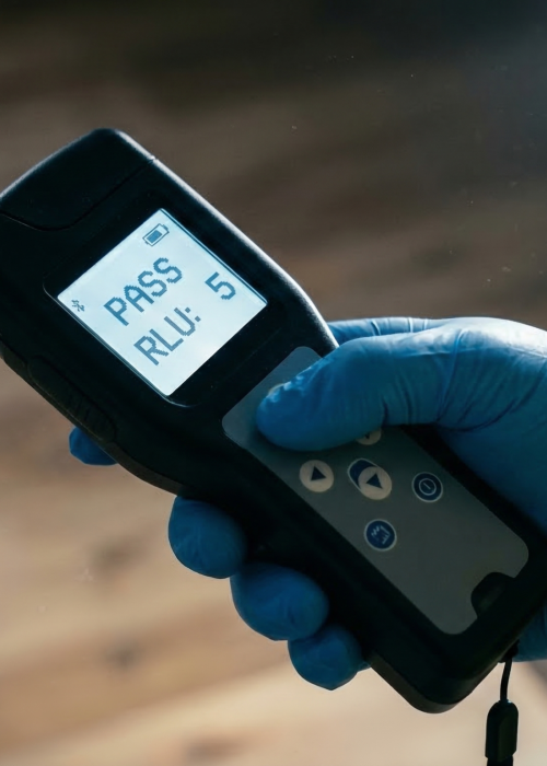 Close-up of an ATP (Adenosine Triphosphate) luminometer held by a blue nitrile glove wearing a Filthy Masters technician in a yellow Tychem hazmat suit, clearly displaying a green 'PASS' result and a score of 'RLU: 5'.